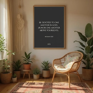 Puede incluir: Una impresión enmarcada con el texto "BE DEVOTED TO ONE ANOTHER IN LOVE. HONOR ONE ANOTHER ABOVE YOURSELVES. ROMANS 12:50". La impresión se exhibe en una habitación con plantas, una silla de mimbre y una alfombra estampada.