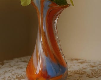 May include: A decorative glass vase with a swirled pattern of orange, blue, and white. The vase holds a bouquet of yellow flowers. The vase is on a table with a floral pattern.