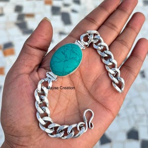 Op de afbeelding: Een zilveren armband met een grote, ovale turkooiskleurige steen in het midden. De armband heeft een chunky kettingontwerp en een haaksluiting. De tekst "Mayae Creation" is zichtbaar op de armband.
