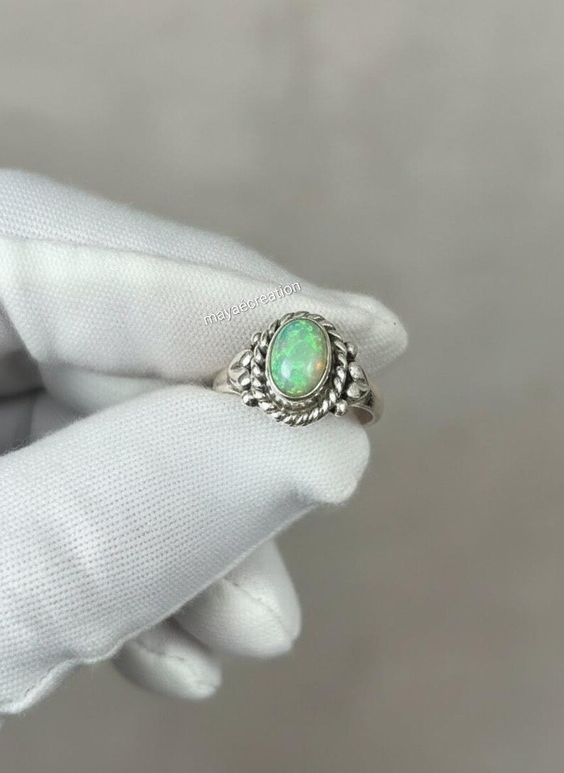 May include: Oval-shaped opal ring with a silver band. The opal has a light green hue and is set in a decorative silver setting with a rope-like border. The ring is held by a gloved hand.