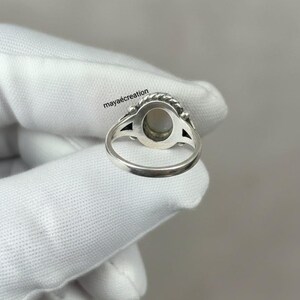 May include: A silver ring with an oval, light-colored stone. The ring has a detailed design around the stone and on the band. The text "maya&eacute;creation" is visible on the image. The ring is held by a gloved hand.