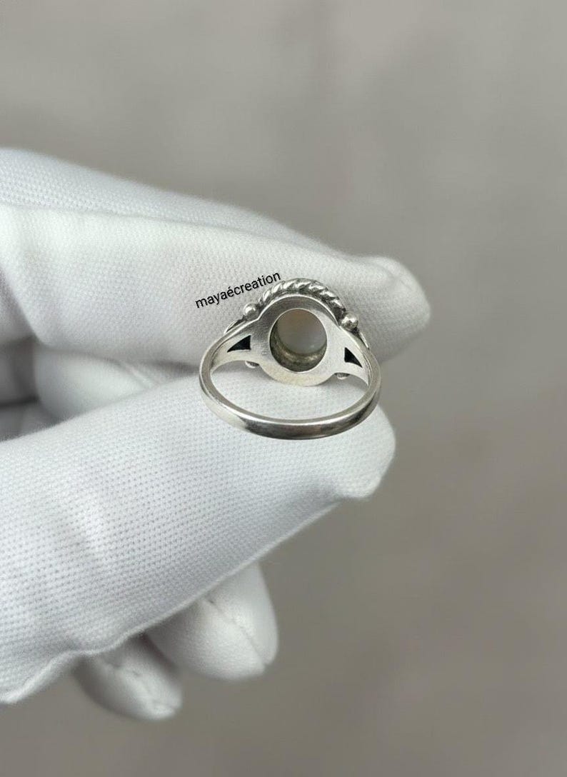 May include: A silver ring with an oval, light-colored stone. The ring has a detailed design around the stone and on the band. The text "maya&eacute;creation" is visible on the image. The ring is held by a gloved hand.