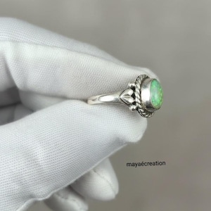 May include: A silver ring with an oval, light green gemstone. The ring has a decorative design around the gemstone and on the band. The ring is held by a gloved hand. The text "mayacreation" is visible.