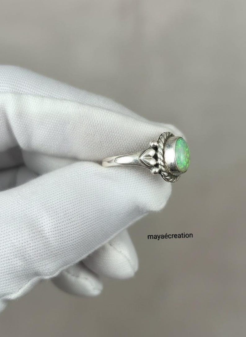 May include: A silver ring with an oval, light green gemstone. The ring has a decorative design around the gemstone and on the band. The ring is held by a gloved hand. The text "mayacreation" is visible.