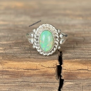 May include: A silver ring with an oval, iridescent opal gemstone. The opal is set in a decorative bezel with a rope-like border. The ring is on a rustic, wooden surface. The text "maya&eacute;creation" is visible.