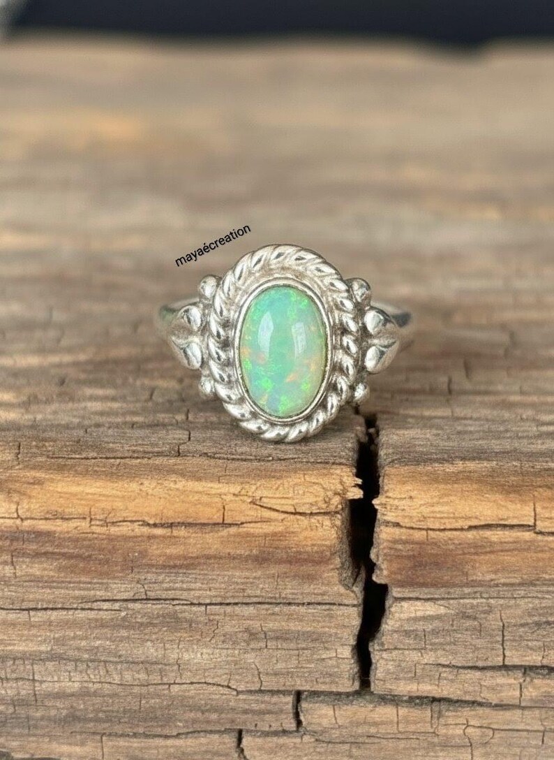 May include: A silver ring with an oval, iridescent opal gemstone. The opal is set in a decorative bezel with a rope-like border. The ring is on a rustic, wooden surface. The text "maya&eacute;creation" is visible.