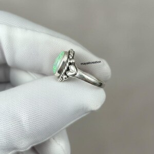 May include: A silver ring with a light green oval gemstone. The ring has a decorative design with small beads and heart-shaped accents. The ring is shown on a white surface.
