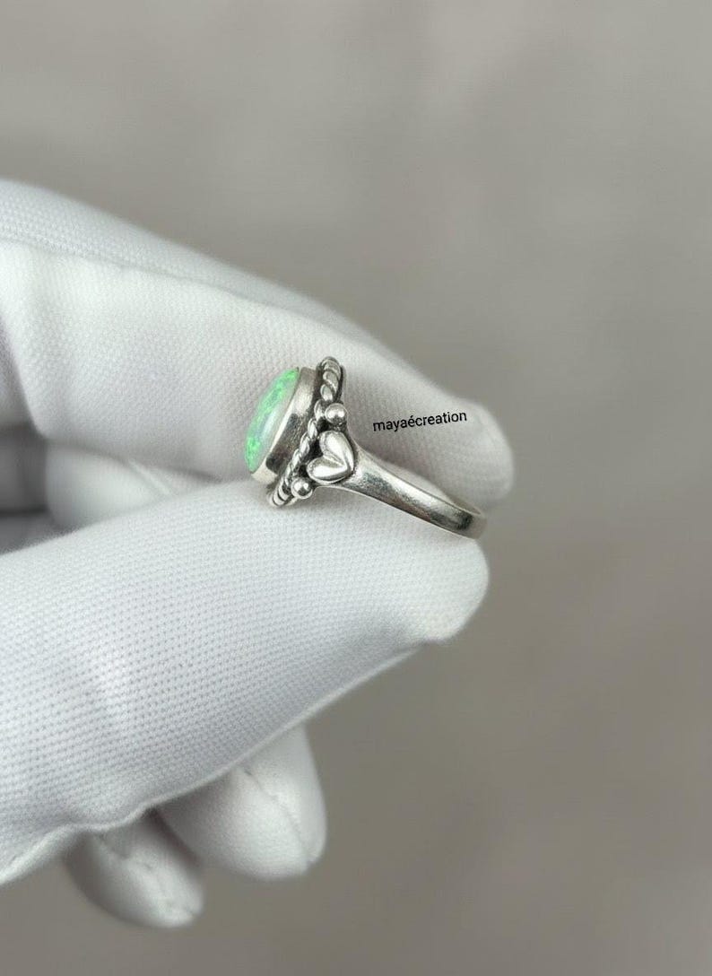 May include: A silver ring with a light green oval gemstone. The ring has a decorative design with small beads and heart-shaped accents. The ring is shown on a white surface.