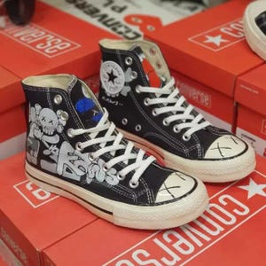 May include: Black high-top sneakers with white laces and a white rubber toe cap. The sneakers feature a KAWS design with cartoon figures in white and gray. The shoes are displayed on a red Converse box.
