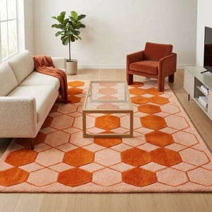 May include: A living room scene featuring a cream-colored sofa, a rust-colored armchair, and a coffee table with a glass top. The focal point is a large area rug with a geometric honeycomb pattern in shades of orange and peach. A potted plant adds a touch of greenery.