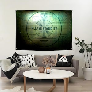 May include: A large, rectangular tapestry with a retro television test pattern design in shades of green and black. The text "PLEASE STAND BY" is prominently displayed. The tapestry is hanging on a white wall above a gray sofa.
