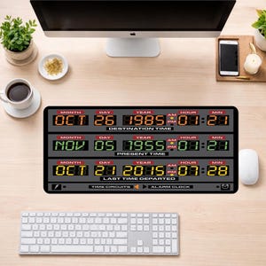May include: A black desk mat featuring a digital time display with the text "DESTINATION TIME", "PRESENT TIME", and "LAST TIME DEPARTED". The display shows dates and times in orange, green, and yellow. A computer, keyboard, and mouse are also visible.