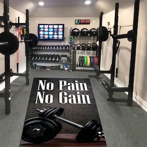May include: A home gym featuring a range of exercise equipment: barbells, dumbbells, kettlebells, and weight plates. A mat on the floor displays the text "No Pain No Gain" with a dumbbell graphic. A digital clock shows the time.