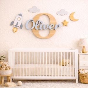 May include: A nursery wall decoration with a wooden letter 'O' and the name 'Oliver' in light blue script. Wooden cutouts of a rocket, clouds, a star, and a crescent moon are also present. A white cot is in the foreground, suggesting a baby's room.