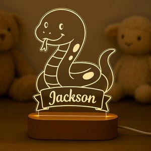 May include: A night light featuring a cartoon snake design with the name "Jackson" on a banner. The light has a warm glow and sits on a wooden base. The snake has a friendly expression.