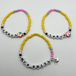 May include: Three beaded bracelets with yellow, pink, and silver beads. One bracelet says "teach" with an apple charm. Another says "learn" with an apple charm. The third bracelet says "mrs smith" with a silver charm.