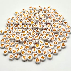 Könnte beinhalten: Ein Haufen weißer, runder Perlen mit orangefarbenen Buchstaben und Zahlen. Die Perlen sind klein, mit einem Loch in der Mitte, und sind auf einer weißen Oberfläche verstreut. Die Buchstaben sind klein.