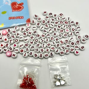 Op de afbeelding: Een verzameling witte, ronde kralen met rode letters en hartvormige kralen. De afbeelding toont ook kleine zakjes met rode en zilveren kralen, en een blauwe kaart met de tekst "LOVE BOMB" en een hart.