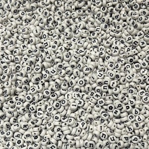 Puede incluir: Una gran colección de pequeñas cuentas redondas de plástico blanco, cada una con una letra minúscula negra o un pequeño símbolo de flor impreso en su superficie. Las cuentas están dispuestas en un montón denso y uniforme, mostrando varias letras.