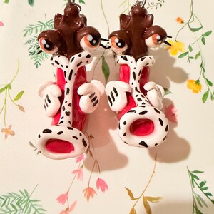 May include: A pair of whimsical, handmade earrings. Each earring features a brown, multi-eyed creature with a red and white body, accented with black spots and white hands. The earrings are set against a floral background.