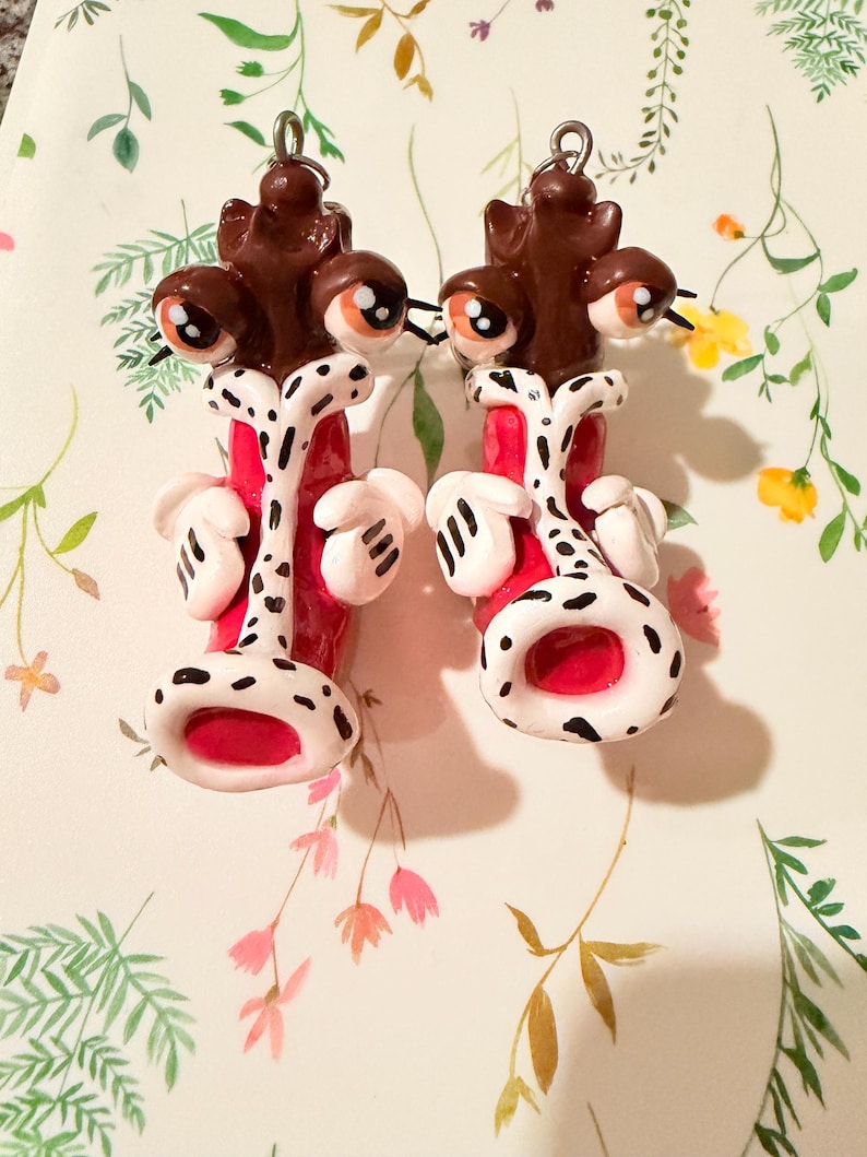 May include: A pair of whimsical, handmade earrings. Each earring features a brown, multi-eyed creature with a red and white body, accented with black spots and white hands. The earrings are set against a floral background.