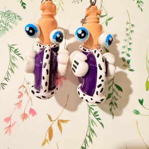 May include: A pair of whimsical, handmade earrings. Each earring features a light orange head with large blue eyes, a purple and white robe, and white gloves. The earrings are suspended from silver-colored hoops.