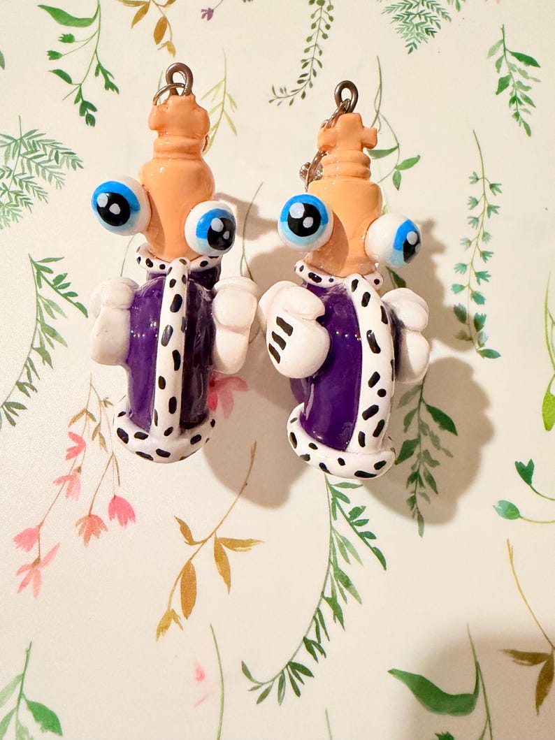 May include: A pair of whimsical, handmade earrings. Each earring features a light orange head with large blue eyes, a purple and white robe, and white gloves. The earrings are suspended from silver-colored hoops.