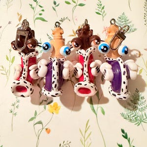 May include: Four whimsical, handmade clay earrings. Each earring features a unique design with large, googly eyes, a red and white body, and a white and black spotted base. The earrings have a playful, cartoonish aesthetic.