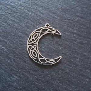 Può includere: Ciondolo a forma di mezzaluna color argento con un intricato design a nodo celtico. Il ciondolo ha un piccolo anello nella parte superiore per appenderlo ed è posto su uno sfondo scuro e strutturato.