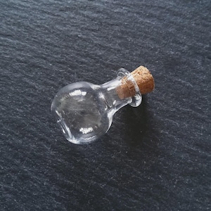 Puede incluir: Una pequeña botella de vidrio transparente con cuerpo redondeado y tapón de corcho. La botella está vacía y descansa sobre una superficie oscura y texturizada. El corcho es de color marrón claro.