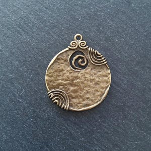 Könnte beinhalten: Bronzefarbener Anhänger mit strukturierter Oberfläche und Spiralmuster in der Mitte. Der Anhänger hat eine runde Form mit dekorativen Wirbeln oben und am Rand, wodurch eine antike oder tribale Ästhetik entsteht.