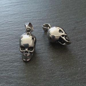Puede incluir: Dos colgantes de calavera plateados con acabado pulido. Los colgantes tienen un lazo para una cadena y detalles faciales. Las calaveras se muestran sobre una superficie gris oscuro.