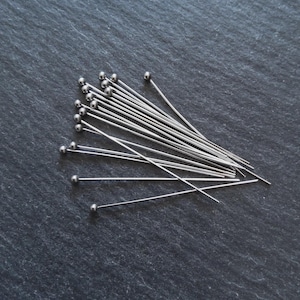 May include: A collection of silver-colored head pins with small, round ball heads. The pins are scattered on a dark gray, textured surface, creating a contrast between the shiny metal and the matte background. These pins are likely used for sewing or crafting.