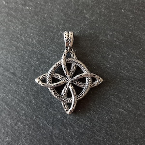 May include: A silver-toned metal pendant featuring a central circular frame with an intricate, interwoven knotwork design that extends into four pointed, petal-like shapes. The surface of the metal has a textured, oxidised finish.
