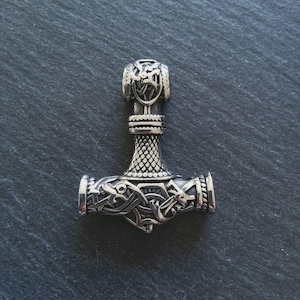 May include: A silver-coloured pendant shaped like Thor's hammer, Mjolnir. The pendant features intricate Viking-style knotwork and design details. The hammer's head and handle are adorned with detailed patterns, and the overall design is symmetrical.