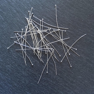 Peut inclure: Un ensemble d'épingles à tête argentées dispersées sur une surface gris foncé. Chaque épingle est surmontée d'une petite boule et d'un long fil droit. Les épingles, de longueurs variables, sont disposées de manière aléatoire.