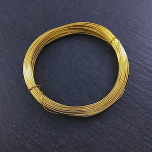 Könnte beinhalten: Eine Spule aus goldfarbenem Draht, eng zu einer kreisförmigen Form gewickelt. Der Draht scheint ein durchgehender Strang zu sein, mit leichtem Glanz. Der Hintergrund ist eine dunkle, strukturierte Oberfläche.