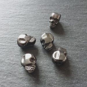 May include: Five small, silver-coloured skull beads are scattered on a grey surface. Each bead is a detailed miniature skull with eye sockets, a nose cavity, and teeth. The skulls are slightly reflective, catching the light.