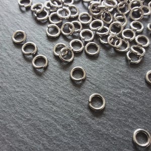 Puede incluir: Una colección de numerosos anillos de salto metálicos circulares de tono plateado. Cada anillo individual presenta un único corte abierto, lo que permite la conexión y el ensamblaje. Los anillos están dispersos por el encuadre.