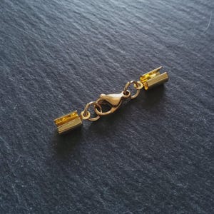 May include: A gold-coloured jewellery clasp with a lobster claw closure. The clasp consists of two rectangular end pieces connected by a small chain and a circular clasp. The background is a dark grey, textured surface.