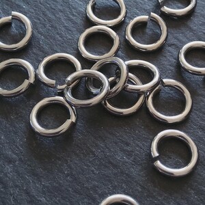 May include: A selection of silver-coloured, open jump rings, arranged on a dark surface. The rings are circular with a small gap, suitable for jewellery making or crafts. The metal has a shiny, reflective finish.