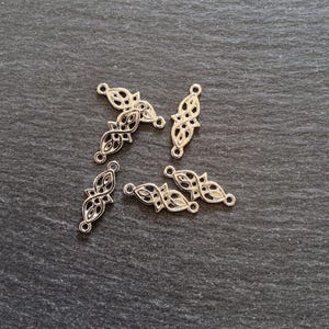 Può includere: Sei ciondoli in filigrana color argento con un design simmetrico. Ogni ciondolo ha due anelli per il fissaggio e un motivo traforato centrale. I ciondoli sono disposti su una superficie strutturata grigio scuro.