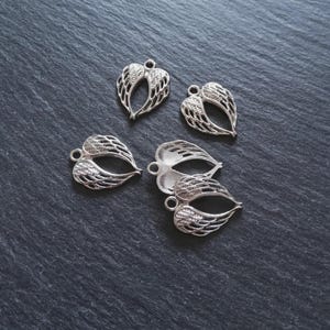 May include: Several silver-coloured heart-shaped charms with wing designs. Each charm has a small loop at the top for attaching to jewellery. The charms are arranged on a dark grey, textured surface.