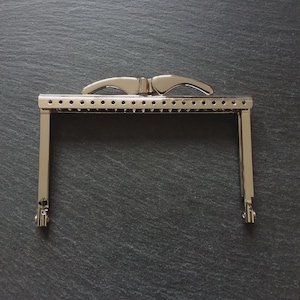 May include: A silver-toned metal purse frame with a rectangular shape and curved handles. The frame has small holes along the top edge for sewing and decorative details. The metal has a shiny, reflective surface.