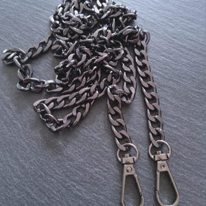 Puede incluir: Una cadena de color metal de ca&ntilde;&oacute;n con eslabones grandes e interconectados. La cadena tiene dos cierres de mosquet&oacute;n en los extremos. La cadena es probablemente para un bolso de mano o un bolso.