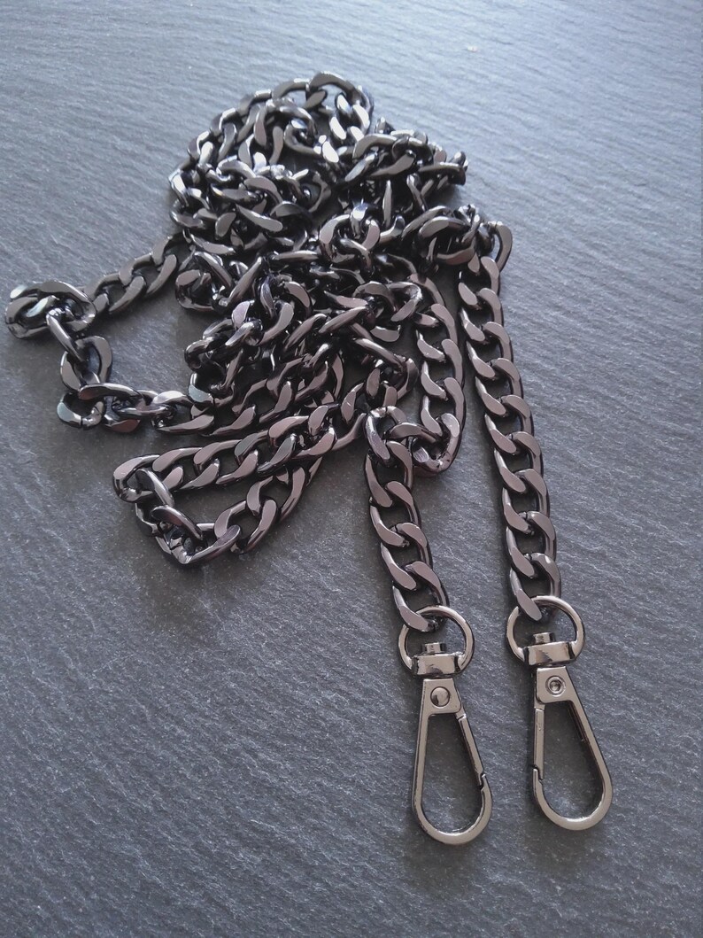 Puede incluir: Una cadena de color metal de ca&ntilde;&oacute;n con eslabones grandes e interconectados. La cadena tiene dos cierres de mosquet&oacute;n en los extremos. La cadena es probablemente para un bolso de mano o un bolso.
