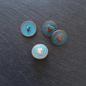 Puede incluir: Cuatro botones decorativos con un aspecto envejecido y antiguo. Los botones son redondos, con una combinación de colores turquesa y cobre. Dos botones presentan un diseño de corazón, mientras que los otros tienen un color turquesa sólido. Los botones son probablemente para coser o manualidades.