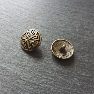 May include: Two antique brass-coloured buttons. One button features a Celtic knot design, while the other shows the back with a central post for sewing. The buttons are round and have a slightly aged appearance.