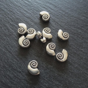 May include: A collection of silver-coloured metal spiral beads, reminiscent of small seashells, are arranged on a dark grey surface. Each bead features a detailed, textured spiral design. These beads are suitable for jewellery making and other craft projects.