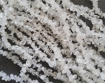 May include: Close-up of multiple strands of clear quartz chip beads. The beads are irregular in shape and size, with a translucent, crystalline appearance. The beads are strung together, creating a textured and organic look. The background is a dark, neutral color.